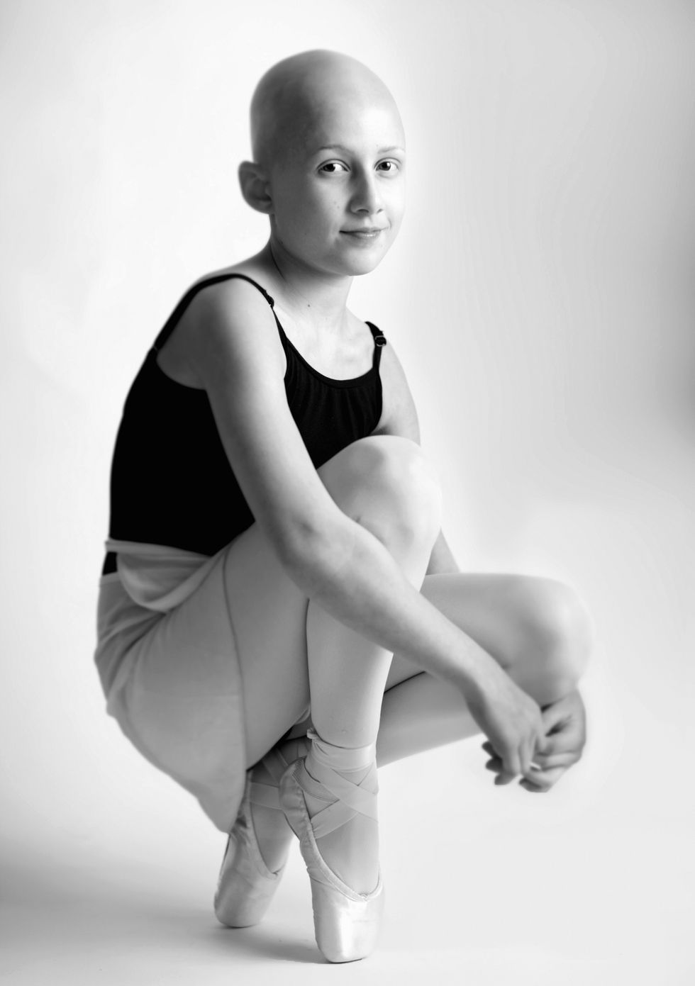 A black and white photo of Stella Jaworski in a leotard, skirt and pointe shoes, crouched down. Stella has no hair.