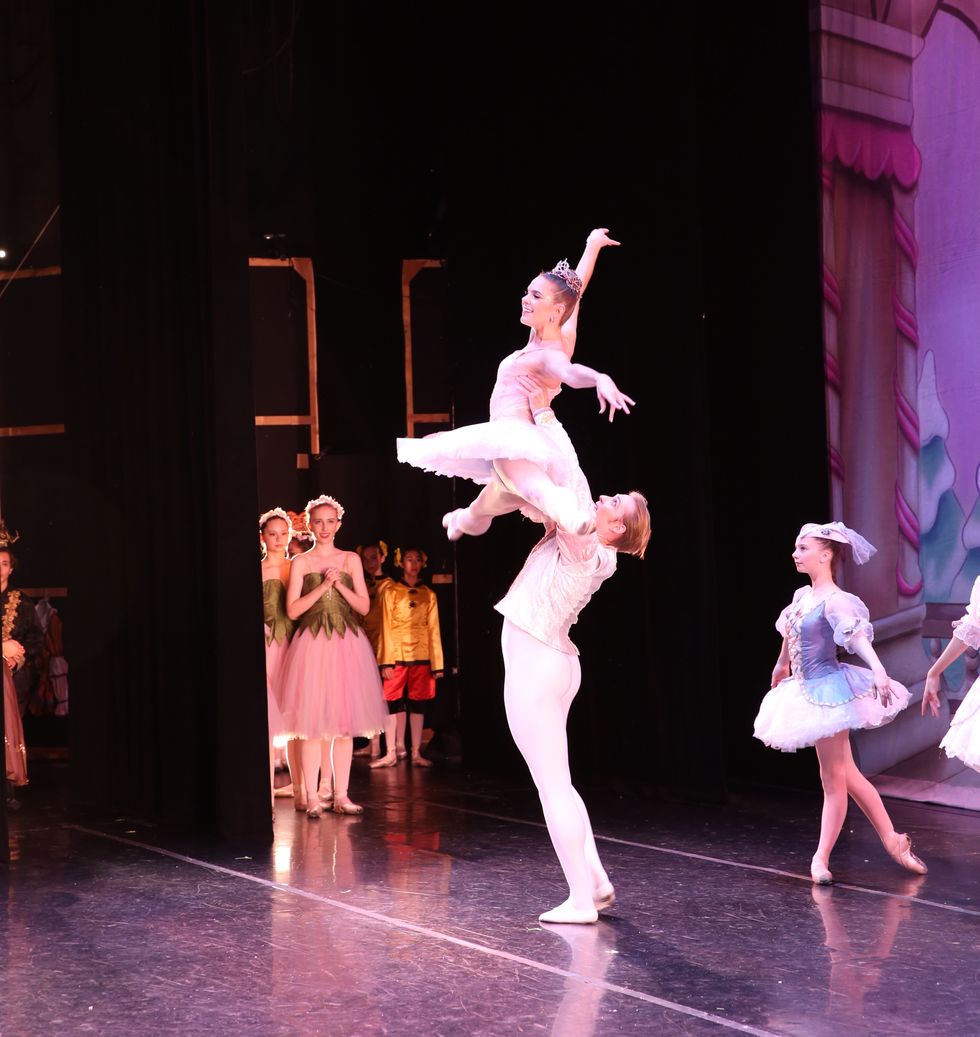 Evan Swensen lifts Savannah Lowrey high into the air during a Nutrcacker performance. She wears a pink tutu, tiara and pointe shoes, he wears a white jacket and tights.