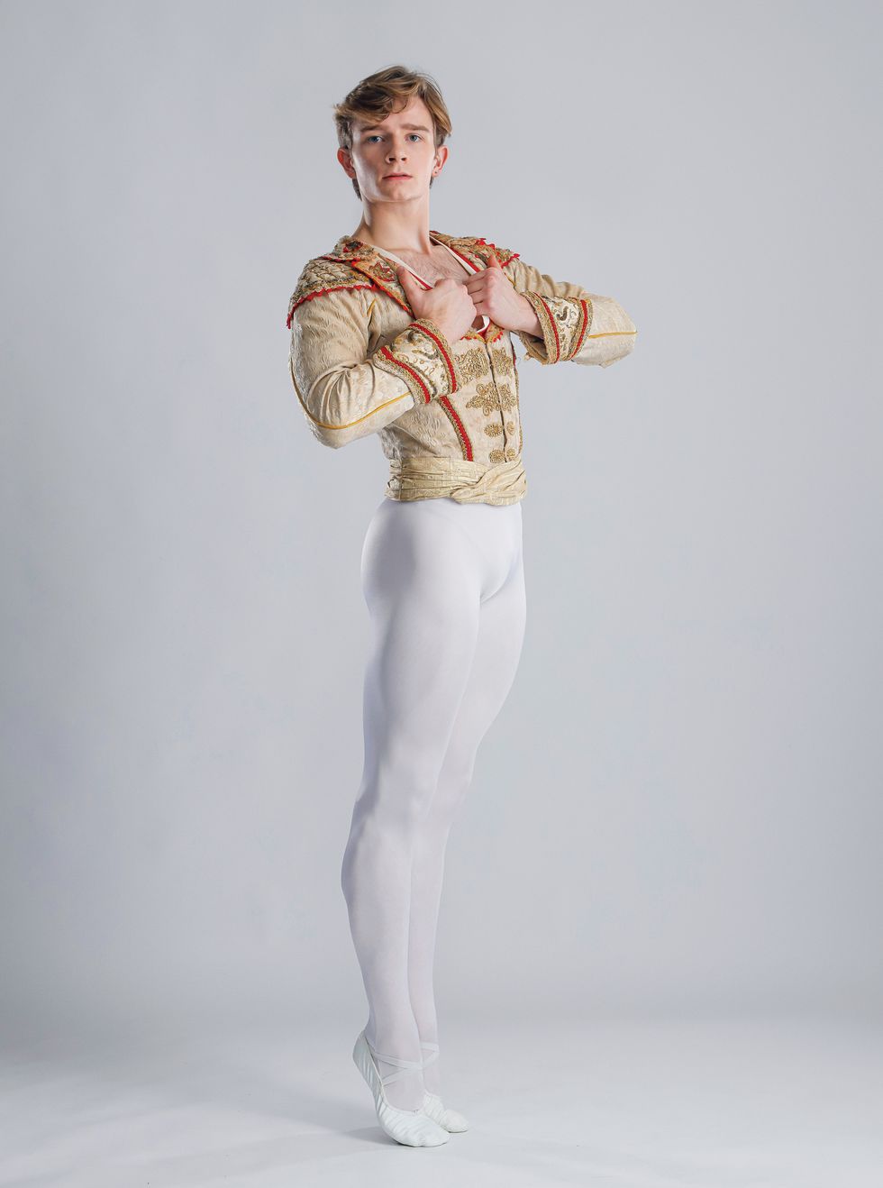 Aran Bell poses against a light grey background. He's wearing white tights and shoes and a gold and Don Quixote jacket.