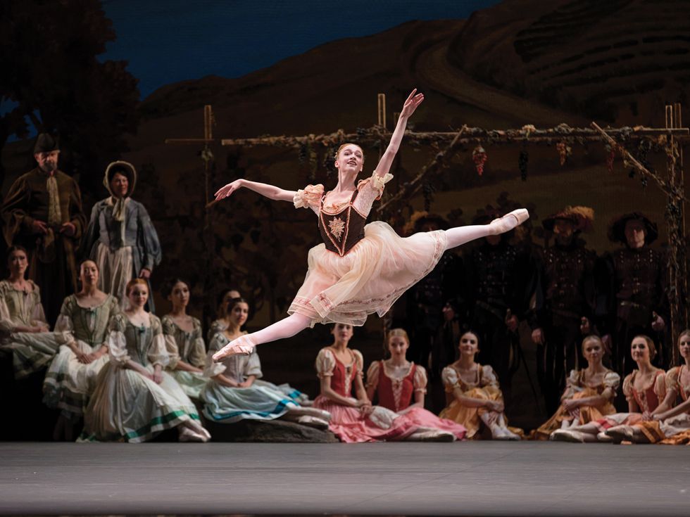 Catherine Hurlin flies through the air in a grand jet\u00e9 wearing a brown and yellow peasant costume. A group of dancers dressed as peasants sit on the ground behind her.