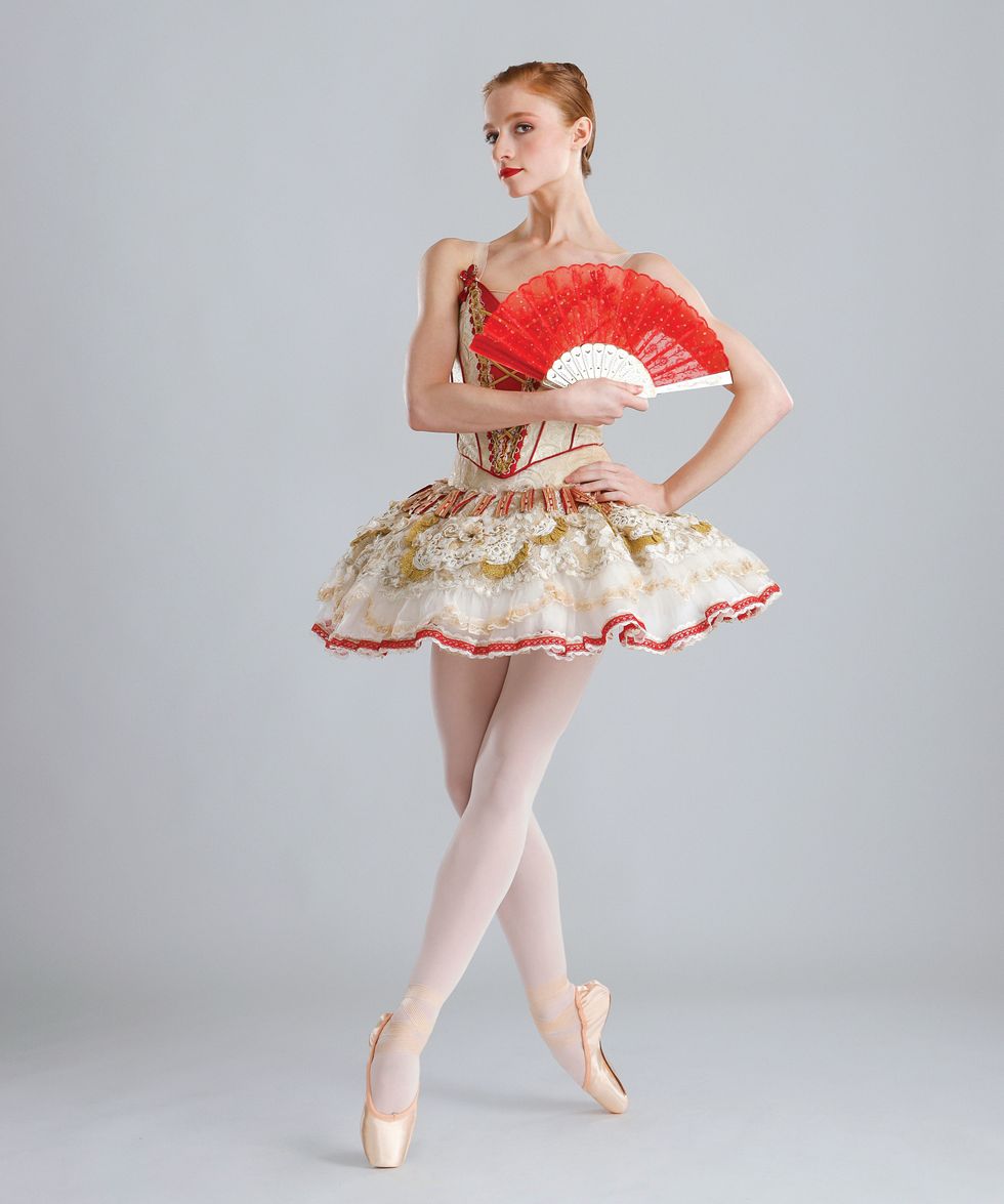 Catherine Hurlin poses with a red fan against a light grey background. She's wearing a red and gold Don Quixote tutu and pointe shoes.