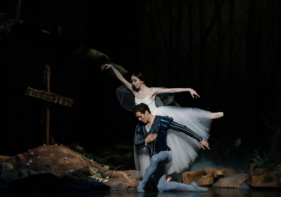 Yuriko Kajiya, wearing a white Romantic tutu, balances in arabesque behind a kneeling Connor Walsh. He wears a dark blue jacket and light blue tights.