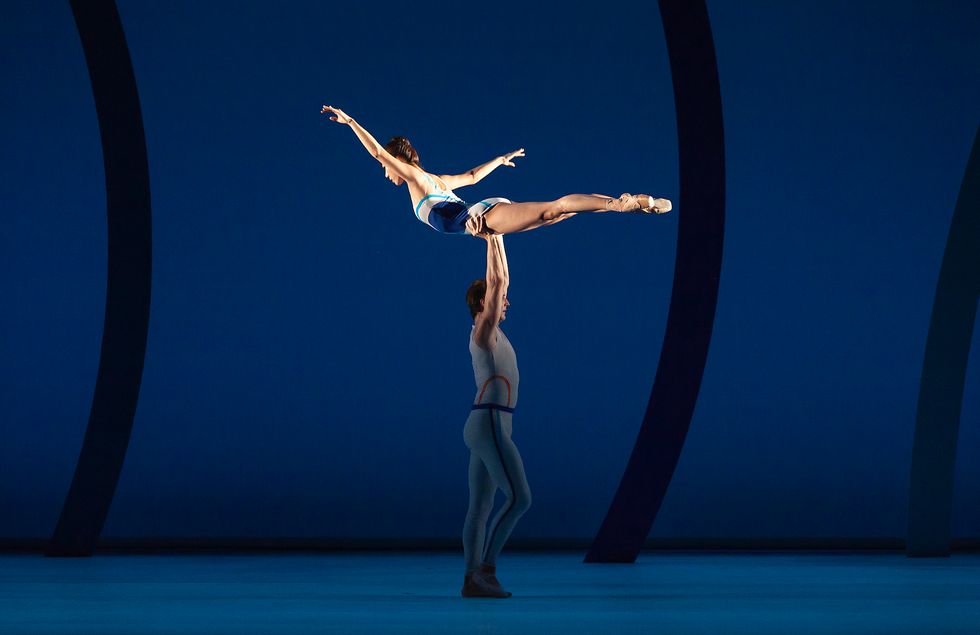 Jacob Bush, wearing a dark blue unitard, lifts Sujin Han, who wears a white and blue leotard, high over his head.