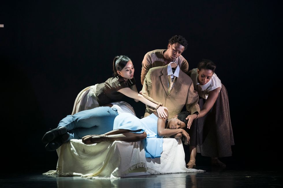 Three dancers hold a brown suit and lean over Cira Robinson as she sleeps onstage.