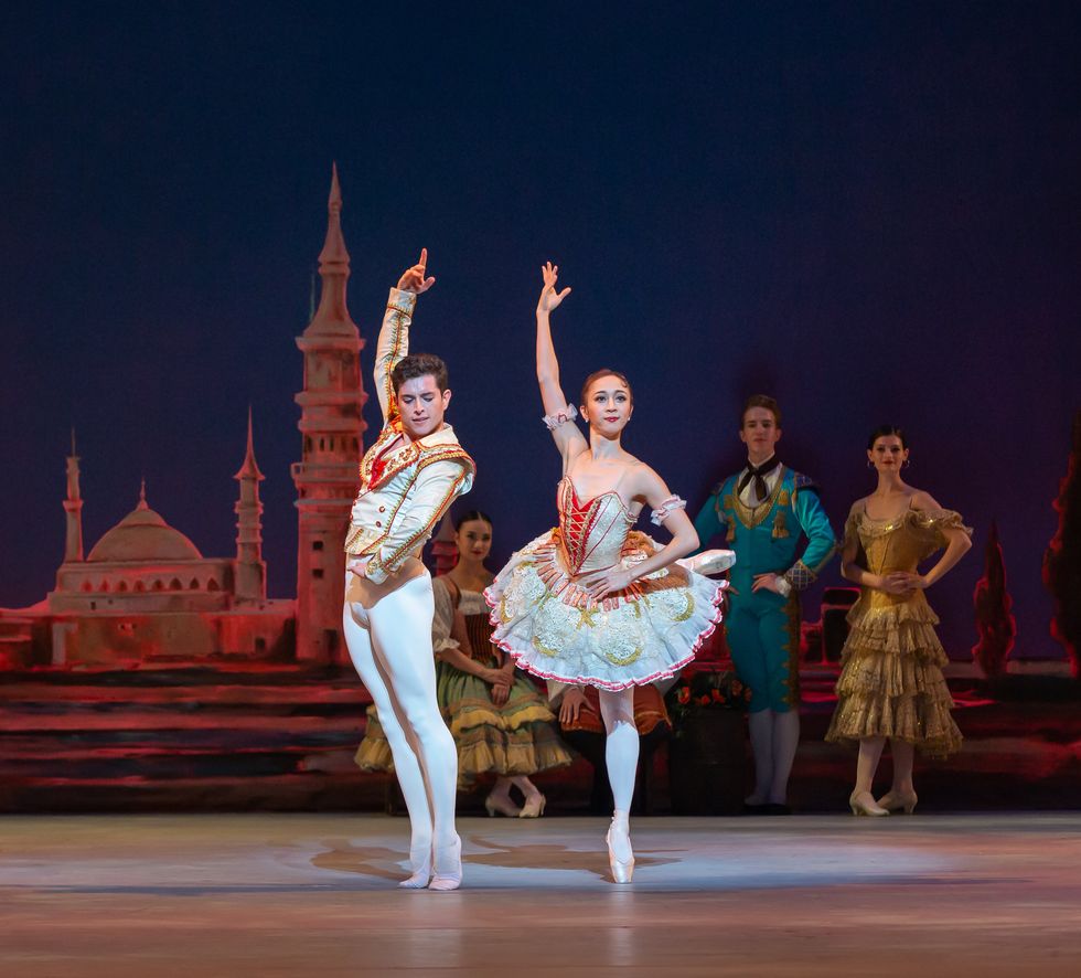 Francisco Estevez and his partner Asuka Sasaki wear white, gold and red Spanish-style costumes. Estevez stands in 6th position on demit pointe while Sasaki balances in attitude derriere.