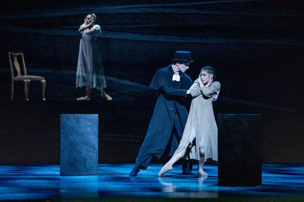 Stefan Goncamvez, wearing a black cloak and top hat, pulls Yumi Kanazawa, who wear a drab brown dress, across the stage. Amanda Assucena covers her ears in the background.