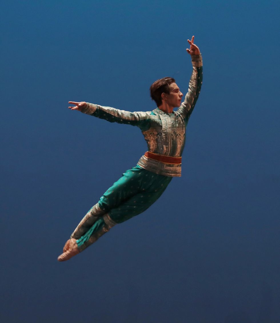 Francesco Mura jumps into the air with his legs pressed togetherin fifth position. His costume includes green and gold tunic and pants.
