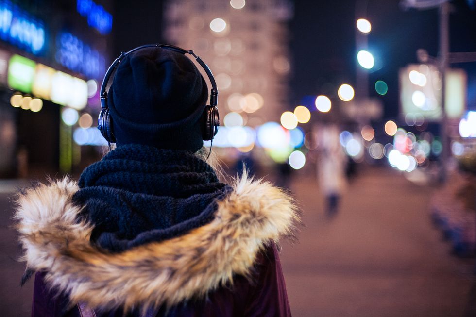 A woman wearing a winter coat, hat and scarf and headphones walks through a city street at night. We only see her from the back.