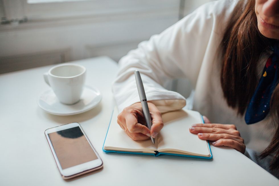 A woman writes in a blue journal on a white table. She wears a white top and has an iPhone and white mug next to her.