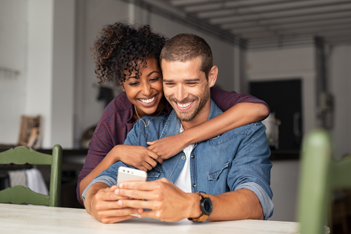 A woman hugs her man who smiles at his phone