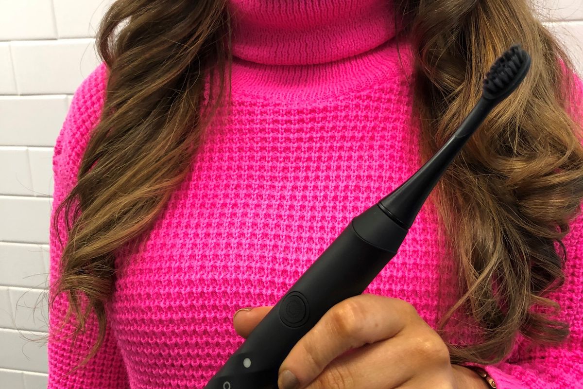 A woman in a bright pink sweater holds up a black BURST toothbrush