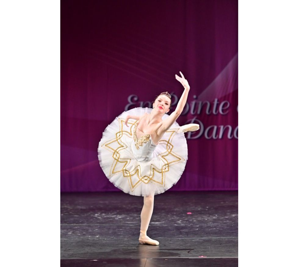 Olivia Book, wearing a white tutu with gold trim, does a renvers\u00e9 in attitude with her right leg onstage during a ballet competition. She looks out to the audeince confidently, with her left arm stretched high above her and her right arm extended to the side.