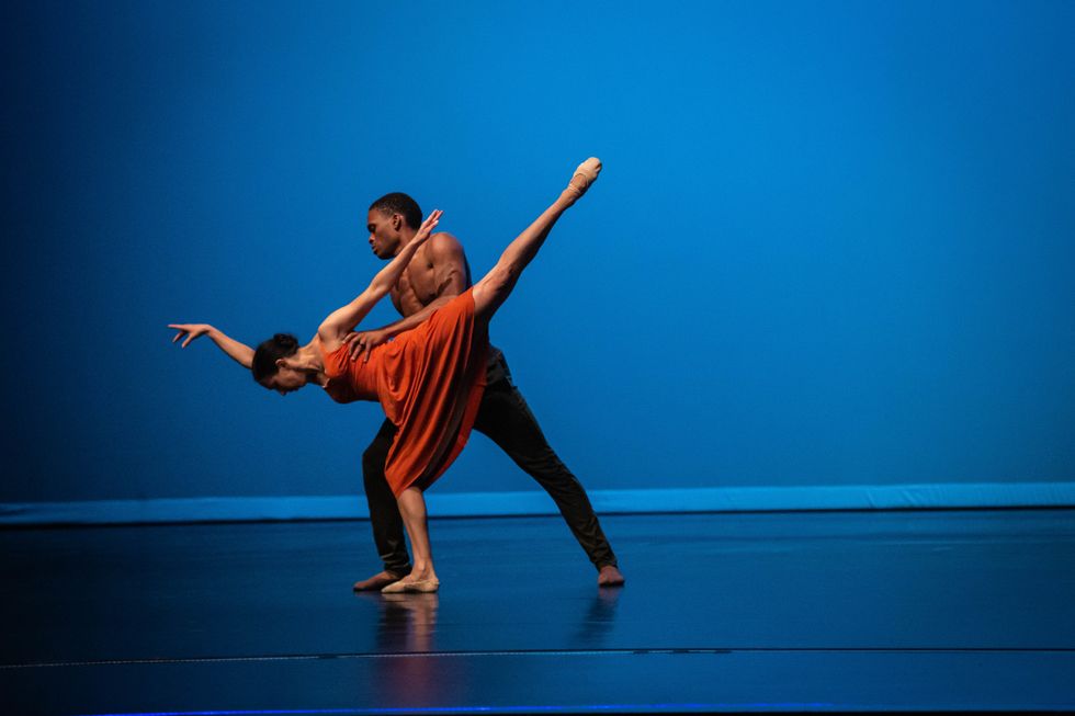 Hill is in a high arabesque pench\u00e9 with her face parallel to the floor. She wears an orange dress and ballet slippers, and is held under her armpits by Vessell, who is standing in a lunge and wearing black pants and no shirt.