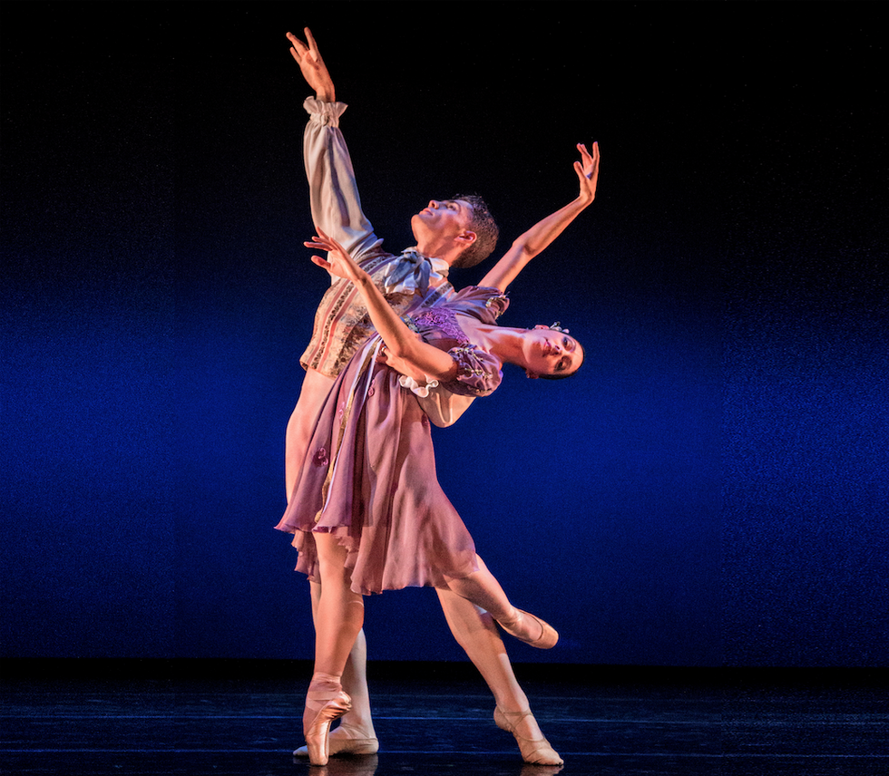 Tilton, dressed in a white princely outfit and standing in a tendu with his arm in the air, holds Farris, who's balanced on pointe in a back attitude in a cambre back. She wears a purple floral dress.