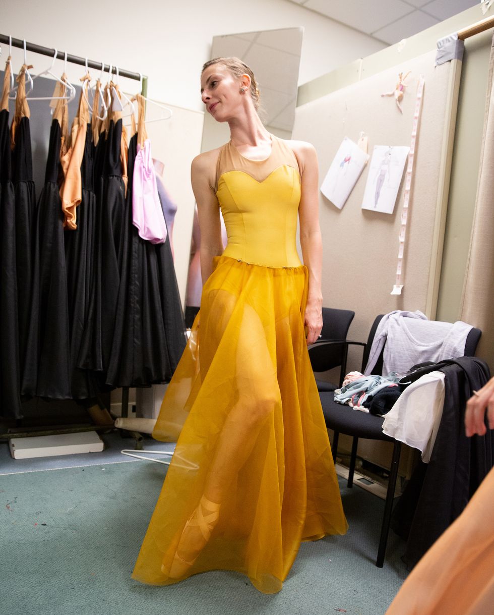 Samantha Hope Galler stands with her right foot popped, looking sideways to the right at her image in the mirror at Miami City Ballet's costume shop. She wears a bright yellow strapless gown; her practice clothes are draped over a black chair behind her.