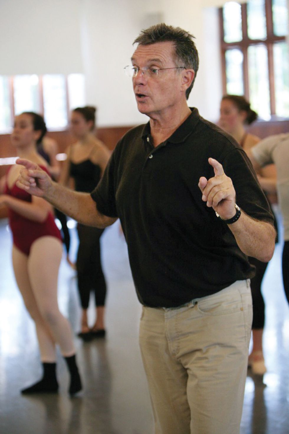 Meehan, dressed in khaki pants and a polo shirt, is in focus in front of a group of ballet dancers in class. He is mid-sentence with his hands in front of him.