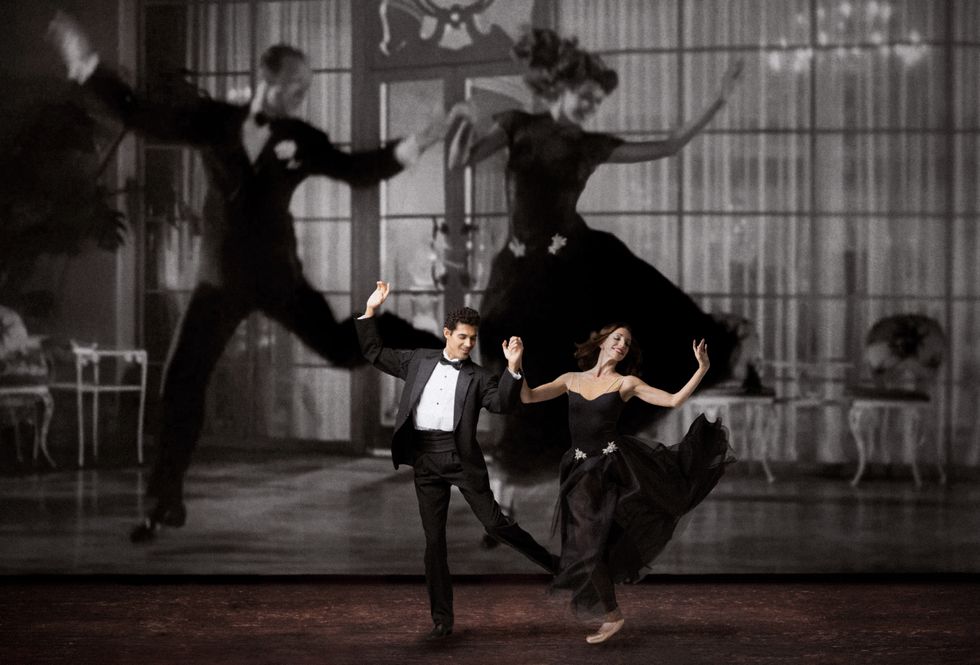 Renan Cerdeiro and Ashley Knox, holding hands, leap lightly onto their right feet, their arms thrown up and their left leg kicked up beside them. He wears a black tuxedo and she wears a strapless black gown. Behind them onstage looms a giant black-and-white projection of Fred Astaire and Rita Hayworth, wearing similar costumes.