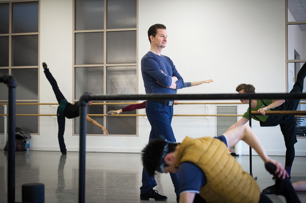 Stark dressed all in blue with his arms crossed, walks through a ballet studio during a men's class. Dancers are stretching at the barre all around him.