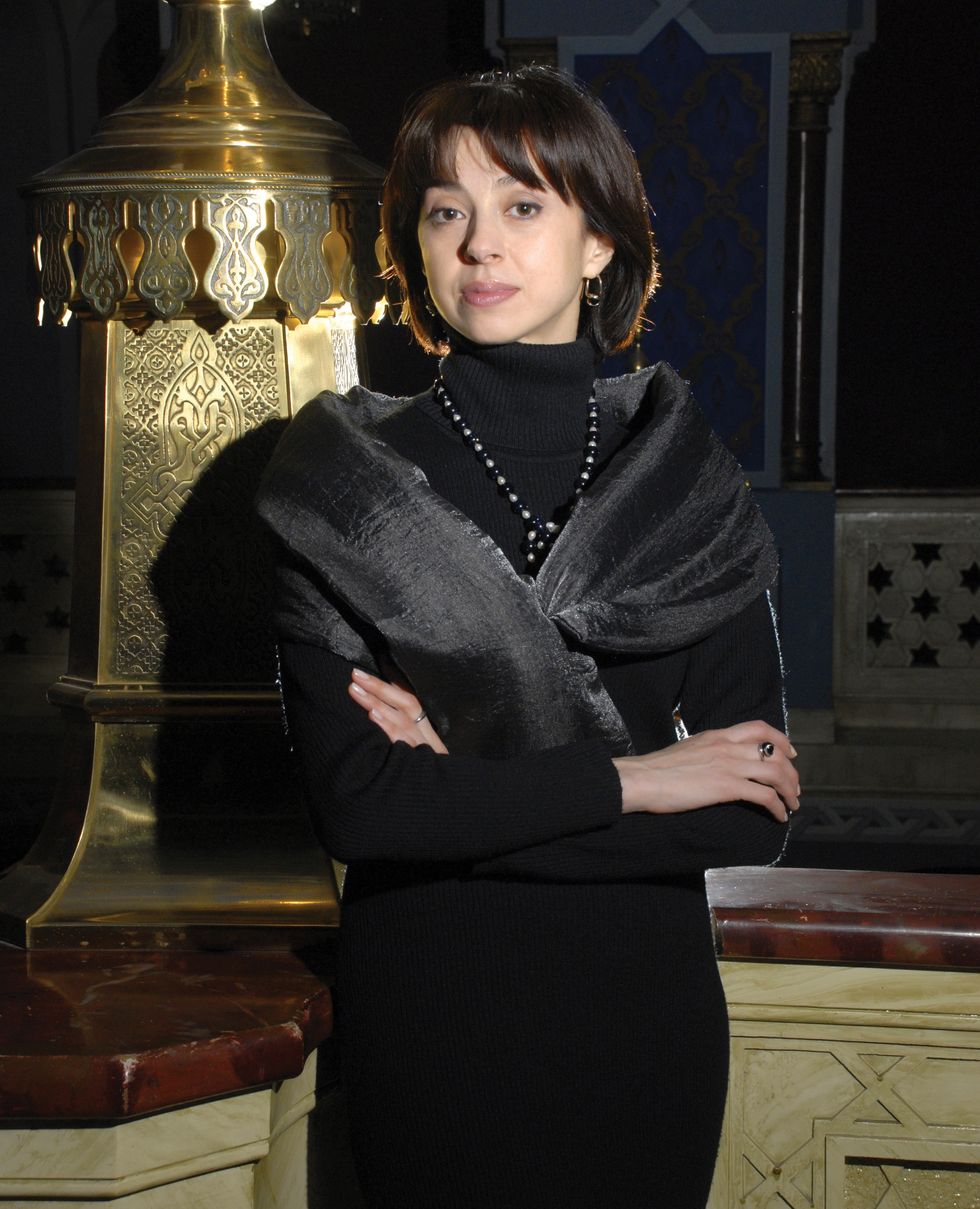 Ananashvili wearing a black dress, scarf and necklace stands in front of a glamorous Russian stage looking background with her arms crossed.