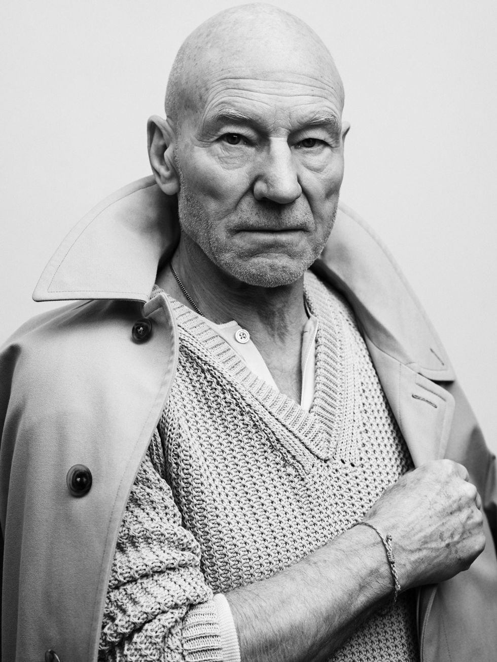 Black and white photo of Sir Patrick Stewart wearing a cable-knit sweater and trenchcoat.
