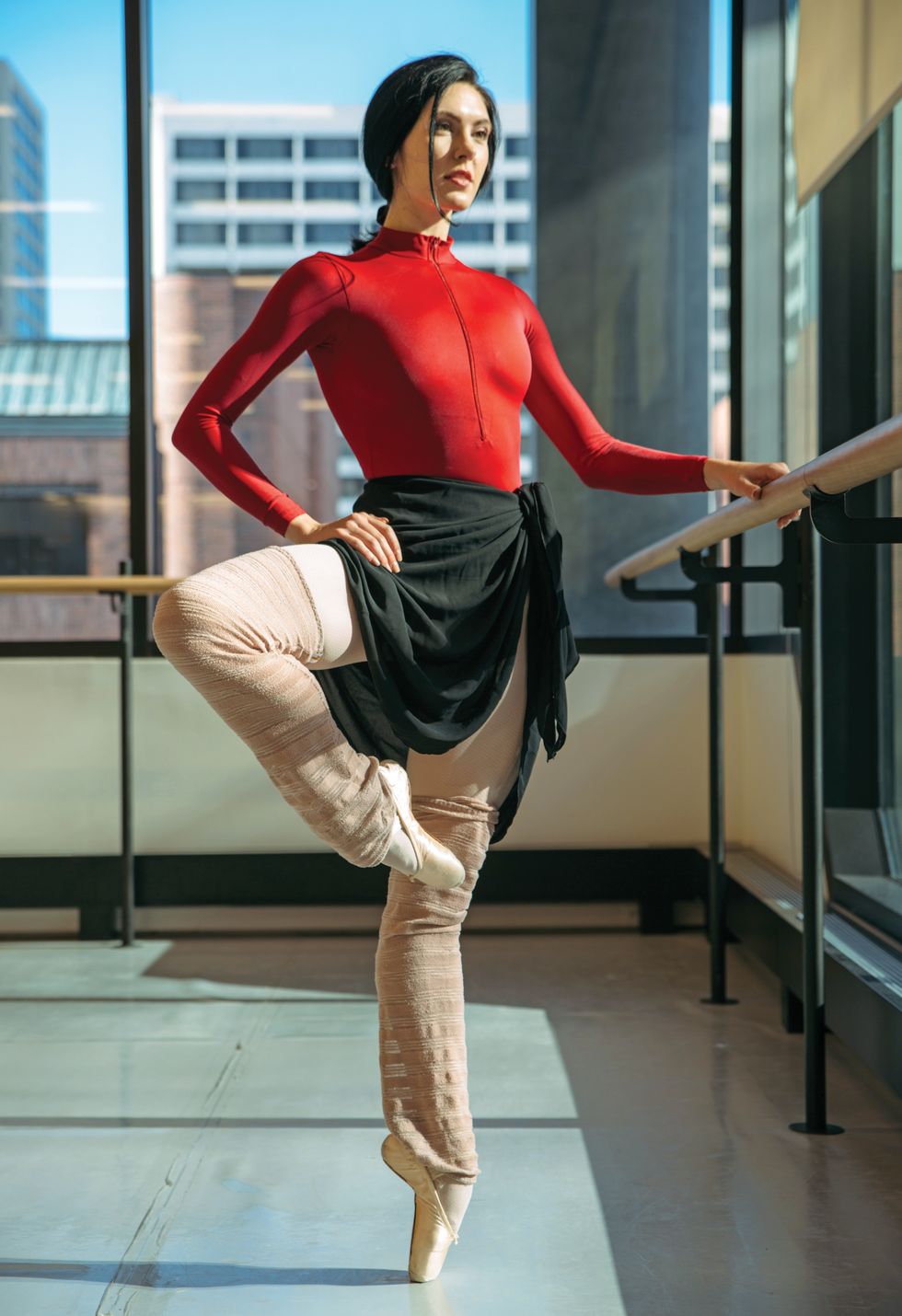 Sisk, dressed in a red leotard and black skirt and legwarmers, stands in a pass\u00e9 on pointe with one hand on the barre.