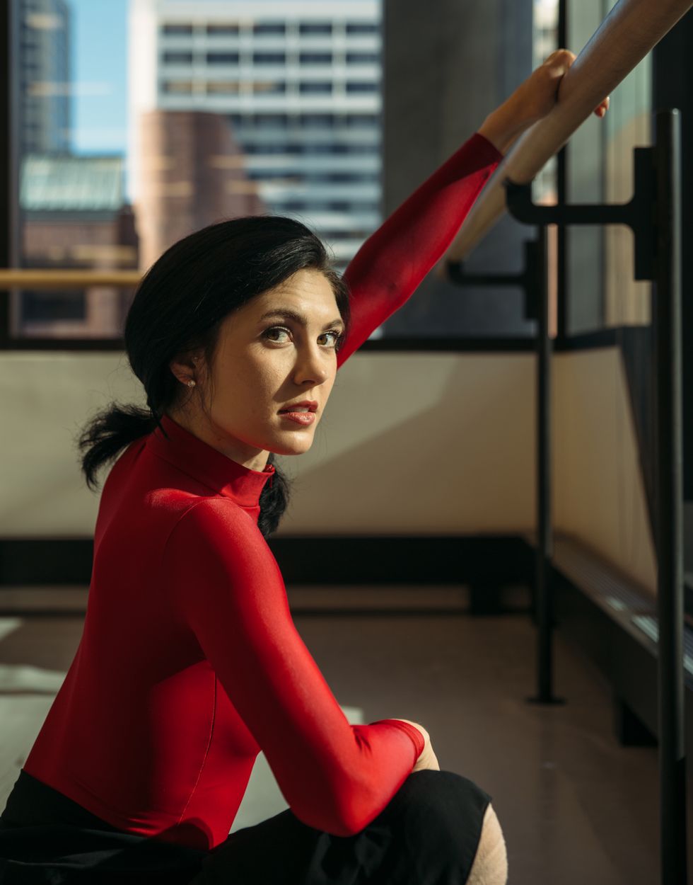 A close-up shot of Sisk holding onto the barre in a shadowy studio with one hand. She's wearing a red leotard and black skirt.