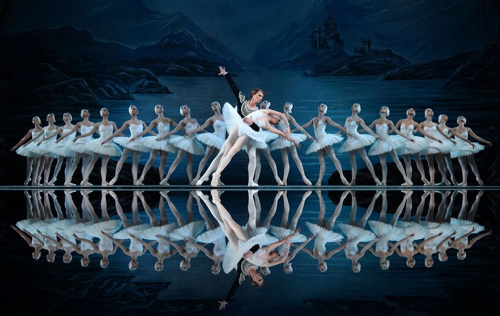 A background of ballerinas dressed as swans frame two dancers. The man, in white tights and a velvet jacket supports the ballerina, in a white swan tutu, who is collapsed over his arm.