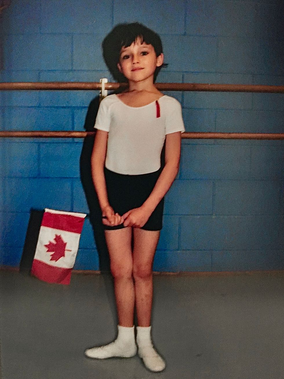 A very young Frenette stands in first position in front of a barre in black shorts, white tee and white shoes with socks. He looks serene and holds a small Canadian flag in his hands.