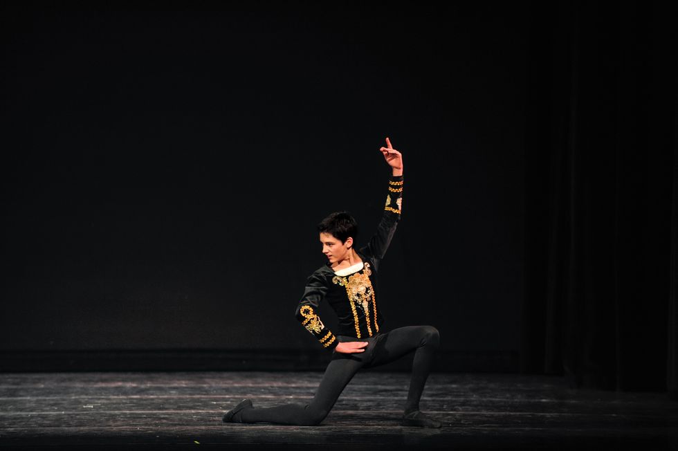 A young Frenette in black tights and a black and gold velvet jacket kneels onstage with one hand on the hip and the other in the air. He looks to the side, as if defiantly at the end of a variation.