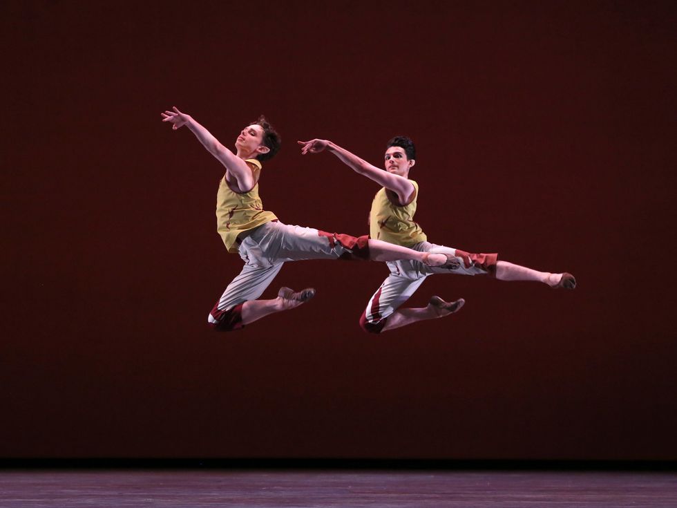 Frenette and Holloway, dressed in patterned grey and red capri pants and yellow and red tops stag leap through the air.