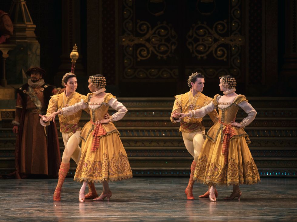 Two couples dressed in elaborate gold and orange outfits stand side by side. In both, the man and the woman look at each other. They both stand in a fourth position and hold hands with the other hand held behind their back.