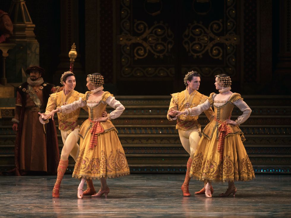 Two couples dressed in elaborate gold and orange outfits stand side by side. In both, the man and the woman look at each other. They both stand in a fourth position and hold hands with the other hand held behind their back.