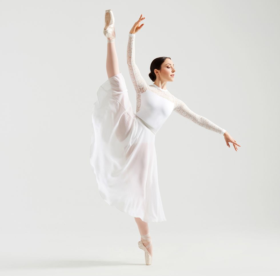 Paraschiv is dressed in a white lace long-sleeved leotard and long white leotard. She's in a high d\u00e9velopp\u00e9 to the site with her gaze over her lower hand.