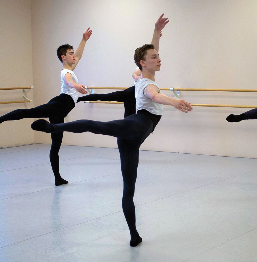 Three boys in black tights and white tee shirts stand in back attitude in a ballet studio.