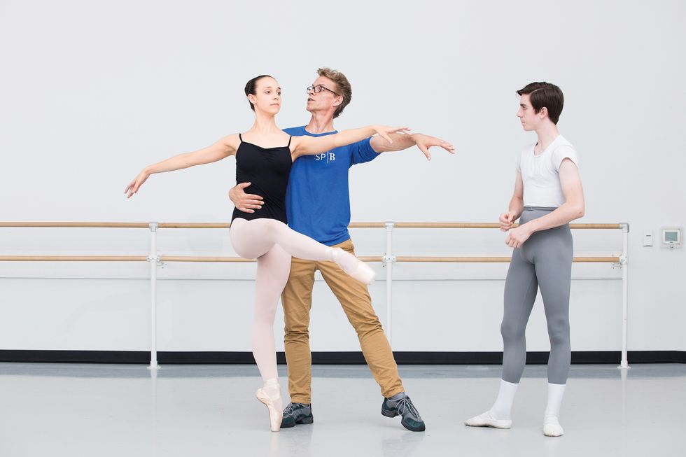 A young ballerina in pink tights and black leotard is being partnered by Payne, while he explains what he's doing. A male student in gray tights and a white tee stands to the side, waiting his turn.