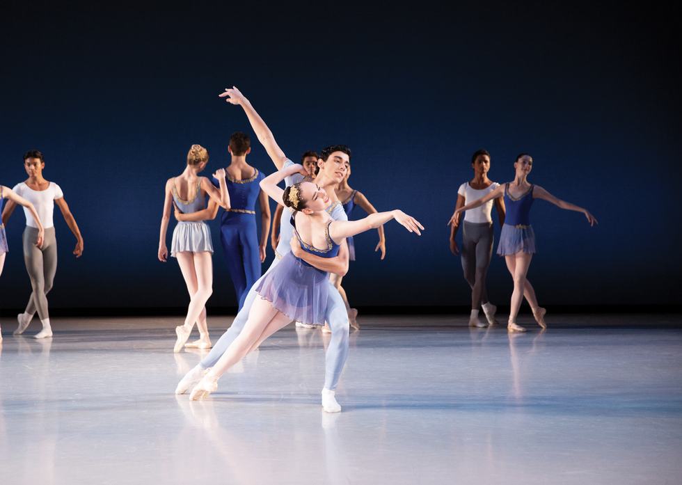 A ballerina in a purplish blue dress and gold hair piece reaches downstage, held by a man in a pale blue outfit. Three other couples stand upstage of them.