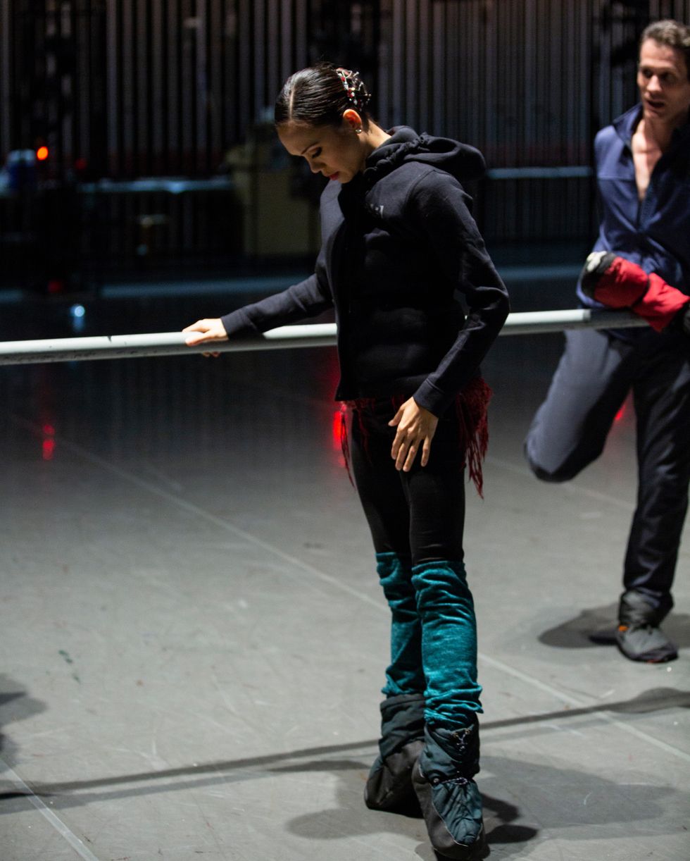 Pantastico, dressed in warm up clothes and booties, stands at the barre in first position relev\u00e9, looking down at her feet.
