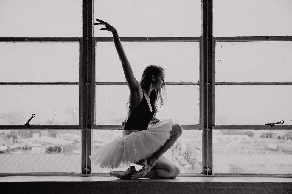 Black and white photograph of Lebowski in a black leotard and white tutu kneeling against a large window in profile. She has one arm lifted.