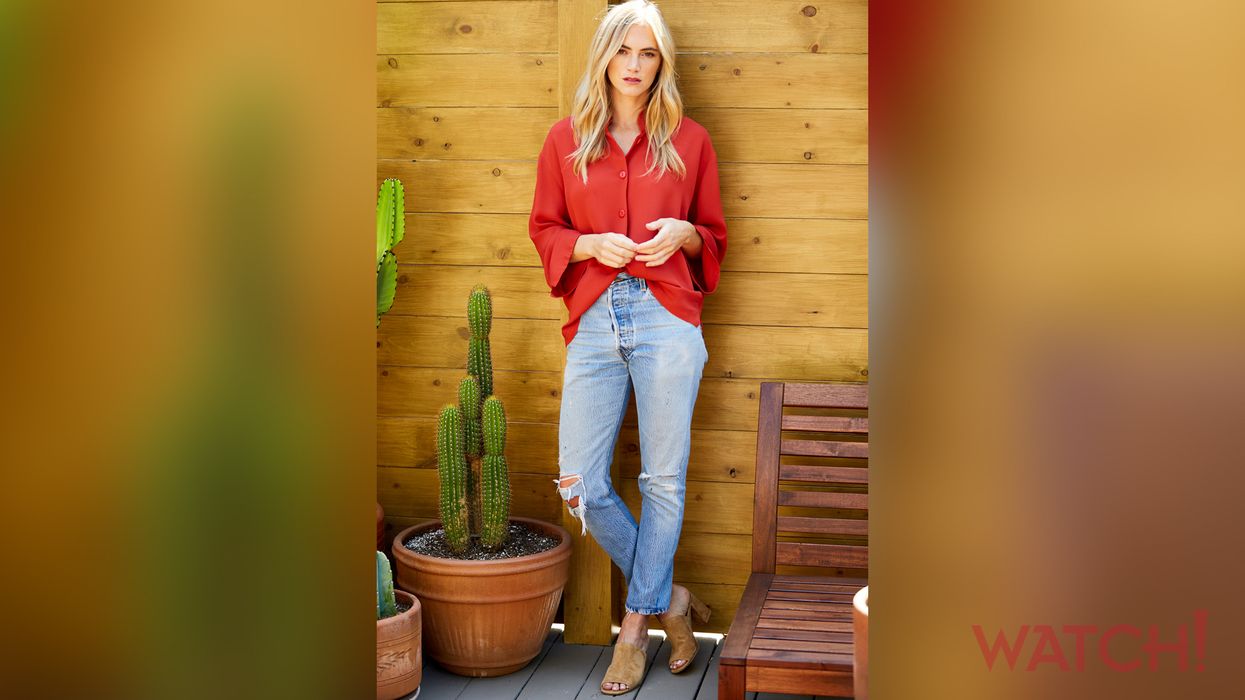 Emily Wickersham leans against a wall wearing a red shirt and ripped blue jeans.
