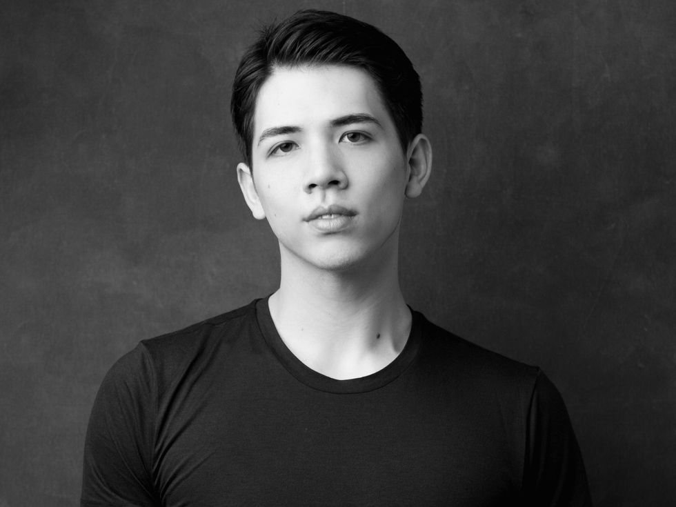 William Yamada is shown from the chest up in a black and white portrait, looking directly at the camera with a small smile. He wears a lack T-shirt and has short dark hair and dark eyes.