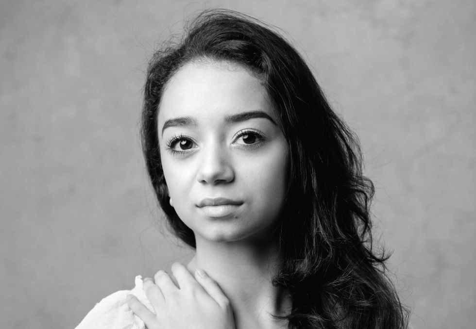 Carolyne Galvao is shown from the shoulders up in a black and white portrait, looking directly at the camera with a small closed smile. She has long, wavy dark hair and wears a white top, and covers her right shoulder with her left hand.