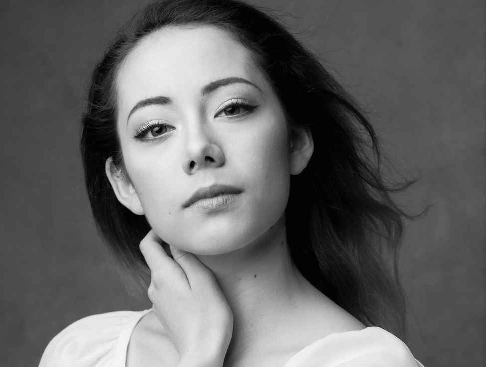 Emily Suzuki is shown from the shoulders up in a black and white portrait, looking directly at the camera with a small closed smile. Wearing a white scoop-neck shirt, she touches her neck with her left hand as her dark hair blows back behind her.