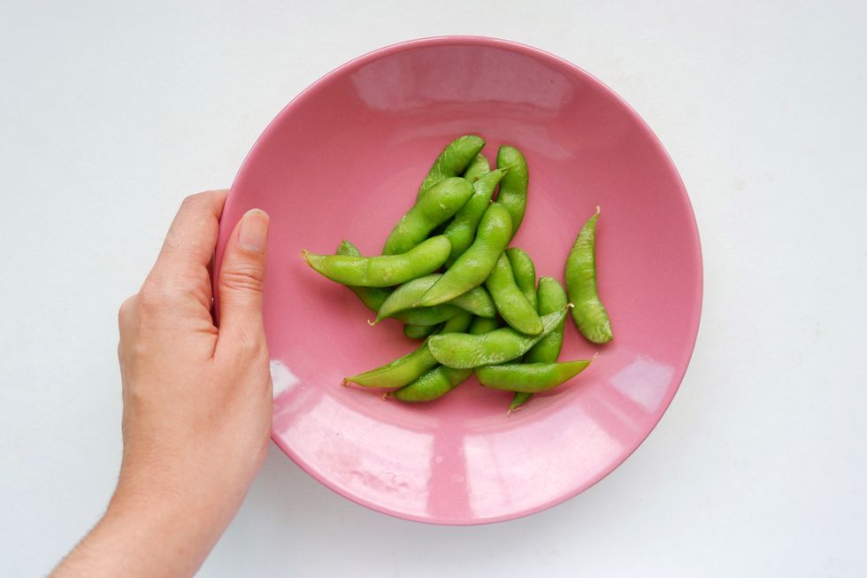 A hand holds a pink bowl filled with edamame, against a white background.