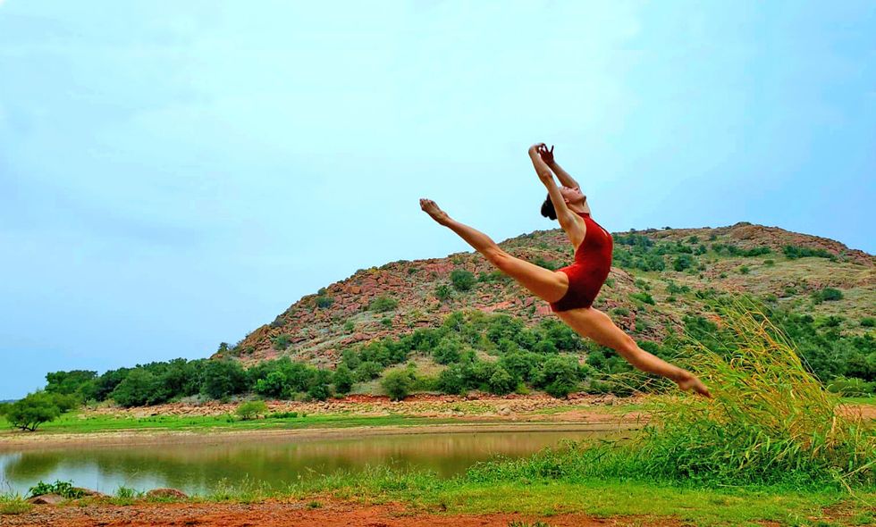 A young woman wearing a red leotard and with bare legs and feet jumps into a powerful sissonne, her legs split and her arms in high fifth, with her head thrown back. She is outside, in front of a small rocky mountain and a body of water, dotted with brush and grasses and under a bright blue sky.