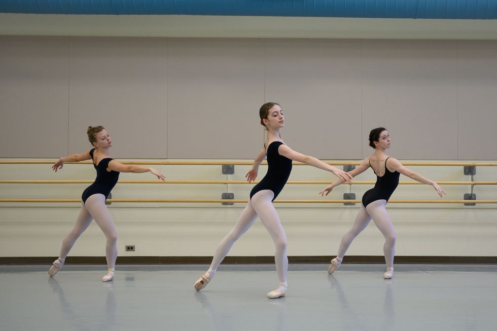 Three ballet dancers take class in black camisole leotards, pink tights and pointe shoes. They are standing in tendu crois\u00e9 derri\u00e8re, with their left leg in pli\u00e9 and their arms in fourth position arabesque.
