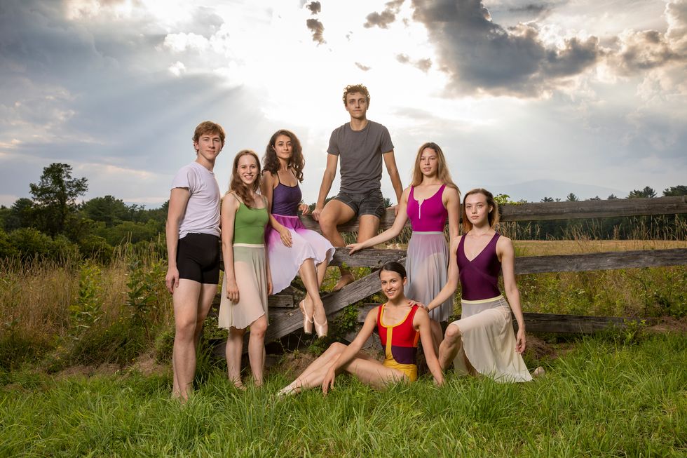 A group of seven ABT Studio Company members pose outside in a field, with two sitting on a gray wooden fence and the rest arranged around them. The two men wear shorts and T-shirts, while the women wear brightly colored leotards and knee-length ballet skirts.