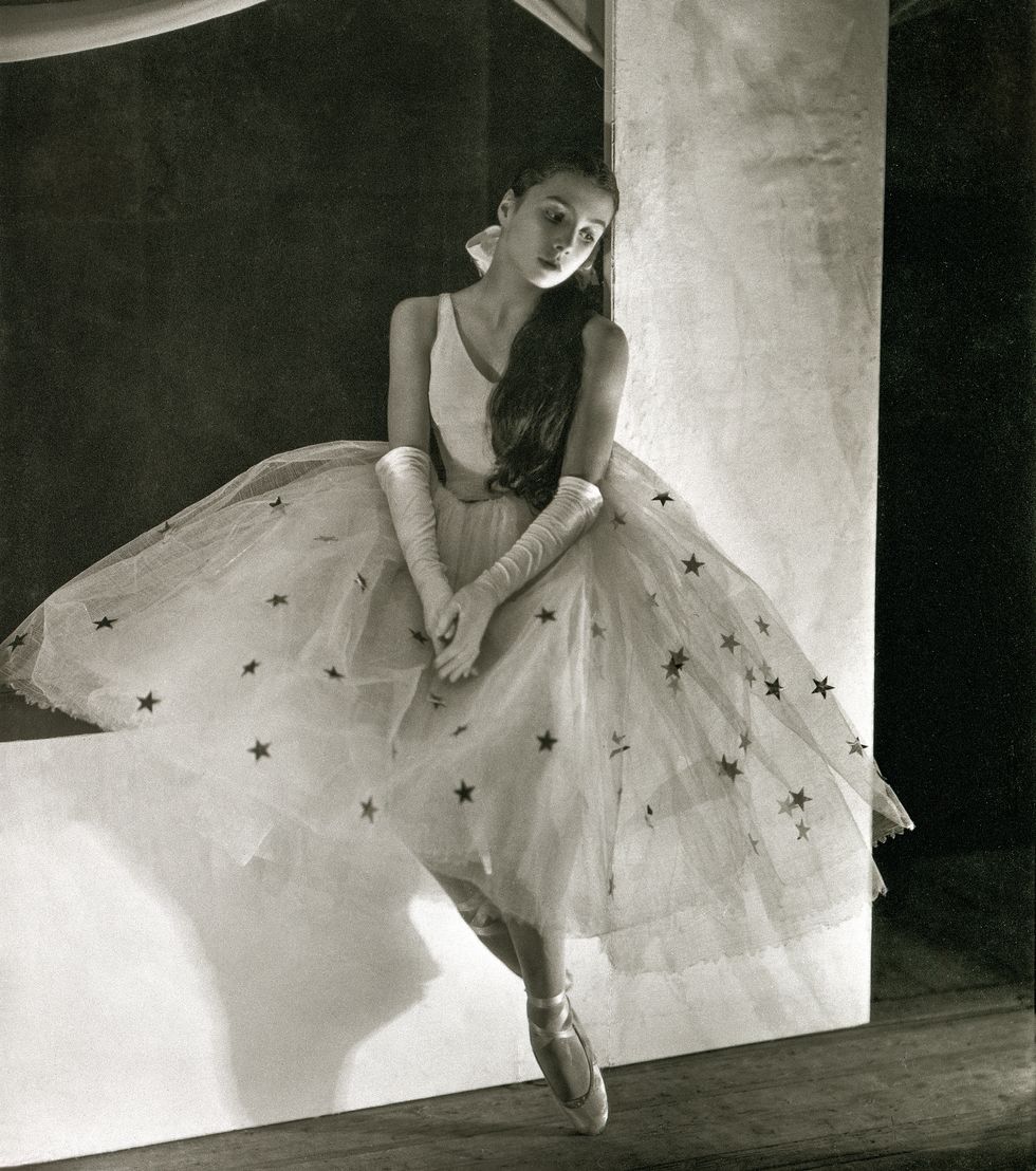 A black and white photo of Toumanova in a large tulle tutu covered in silver stars, white bodice and white gloves sitting on a large white set piece with her hands in her lap.