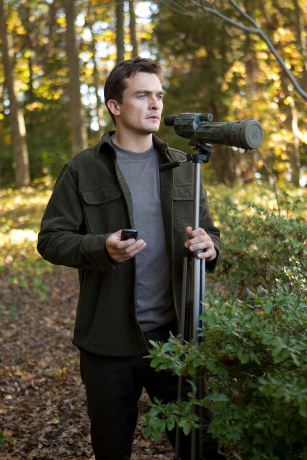 Rupert Friend behind a telescope in the woods.