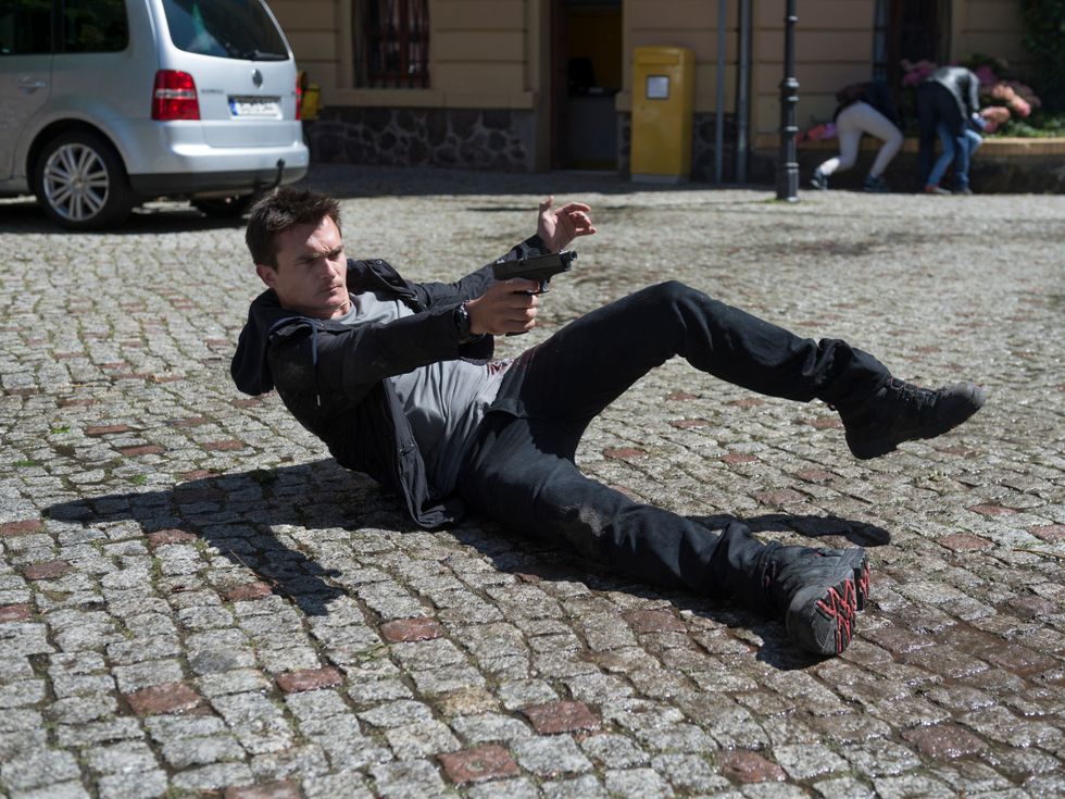 Rupert Friend lying on cobblestone street with a gun