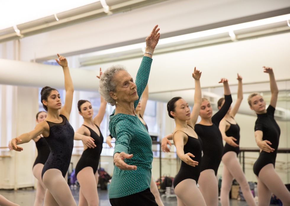 Lupe Serrano, shown from the waist up and wearing a long-sleeved green shirt and black pants, demonstrates tendu crois\u00e9 devant. A group of teenage dancers in lack leotards and pink tights stand behind and to the right of her, imitating her position.